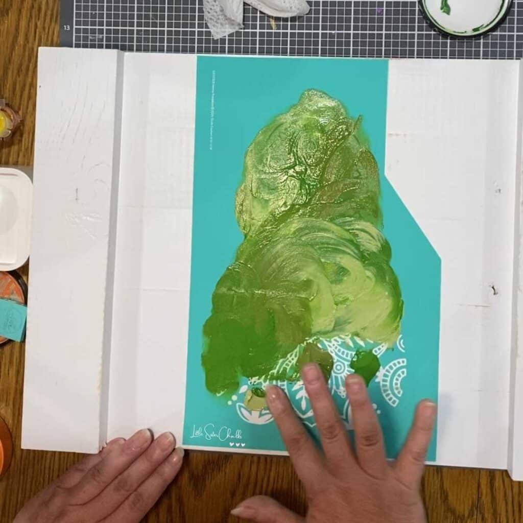 Squash stencil on white wooden board with hand smearing various shades of green paste across stencil for blog post The Marbling Technique with Chalk Couture is a Hot Mess