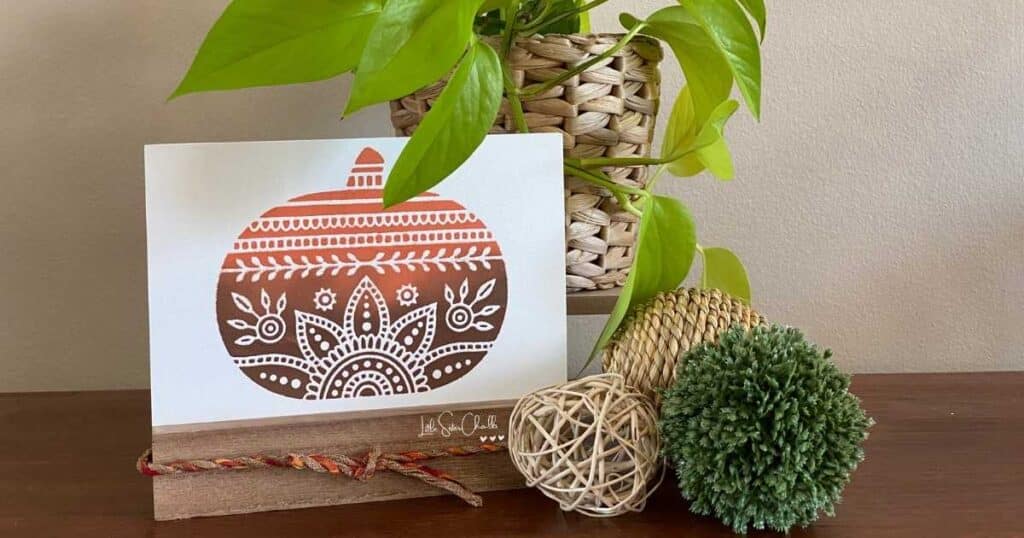 A pumpkin chalked in the ombre effect with orange and brown chalk paste on a white board surface in a brown base with a multi colored ribbon in coordinating colors next to two neutral spheres and one green sphere in front on a neon pothos in a woven container for the blog post The Chalk Couture Ombre Effect