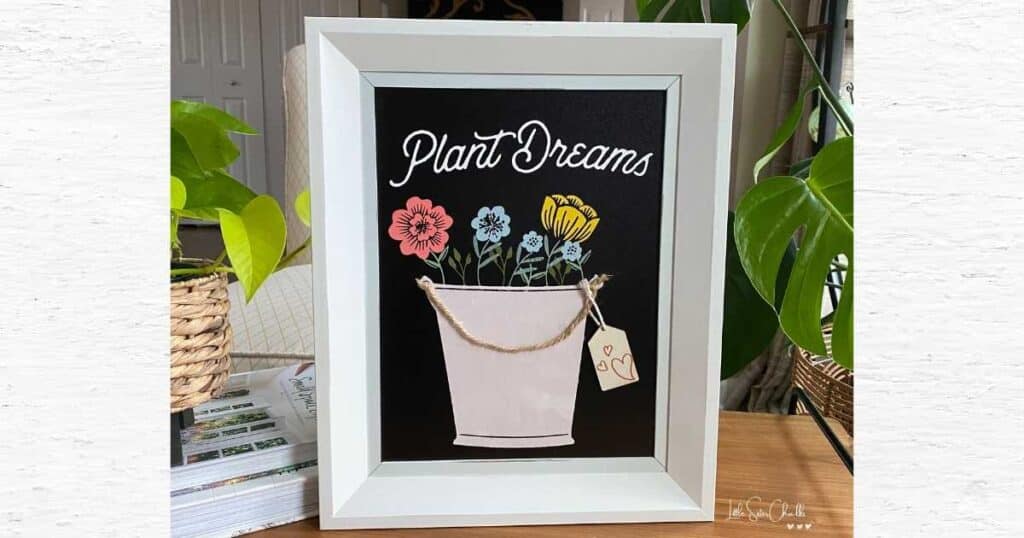A white framed chalkboard surface with a chalked design of a bucket with 5 flowers in coral, blue and yellow with the words "plant dreams" for blog post Layering Chalk Couture Stencils