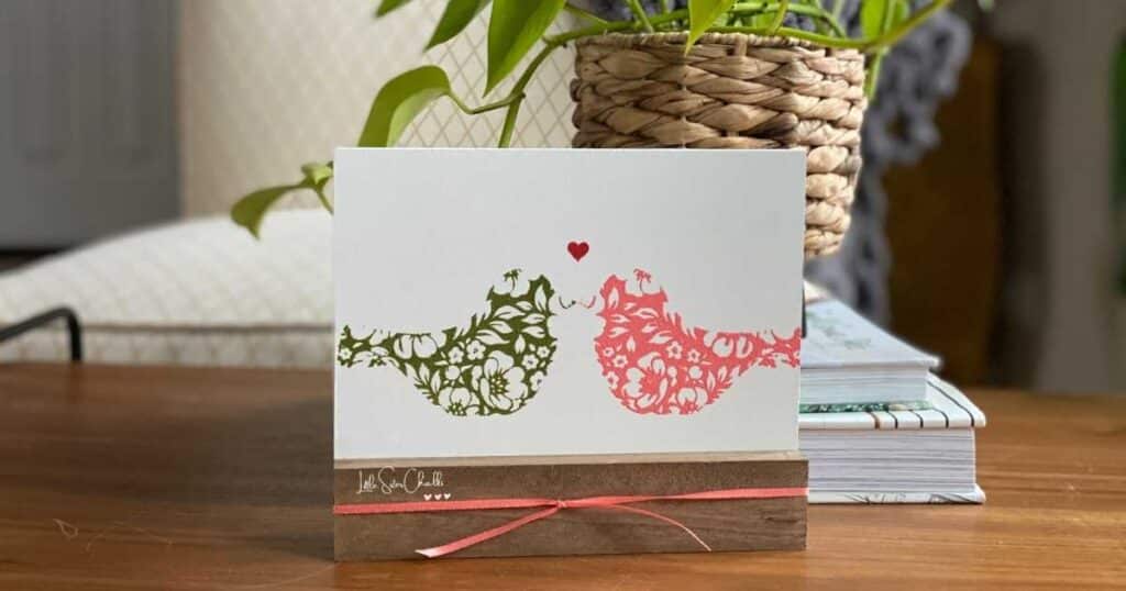Image of a bird chalked in green chalk paste on the left with its mirror image chalked in coral chalk paste on the right on a white board in a brown wood base with a small heart chalked between the birds and coral ribbon around the base in front of a neon pothos in a woven pot all on a coffee table in front of an off-white chair with a gold diamond pattern for the blog post Creating Mirror Images Using Chalk Couture