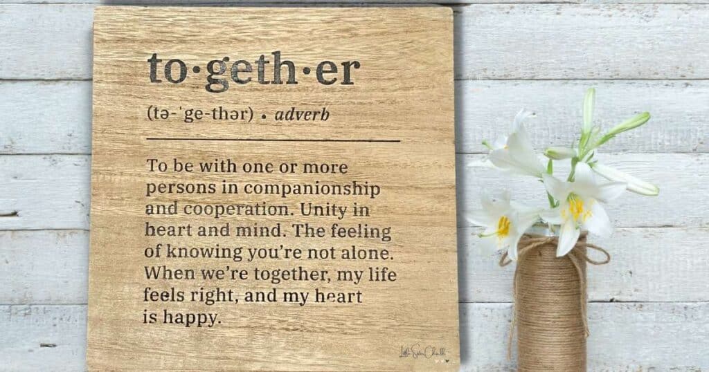 A wooden sign chalked with the words "to-geth-er" with its definition reading "To be with one or more persons in companionship and cooperation. Unity in heart and mind. The feeling of knowing you're not alone. When we're together, my life feels right, and my heart is happy" next to a vase covered with twine  holding white lilies for the blog post How to Quickly Create an Easy One-and-Done Chalk Project