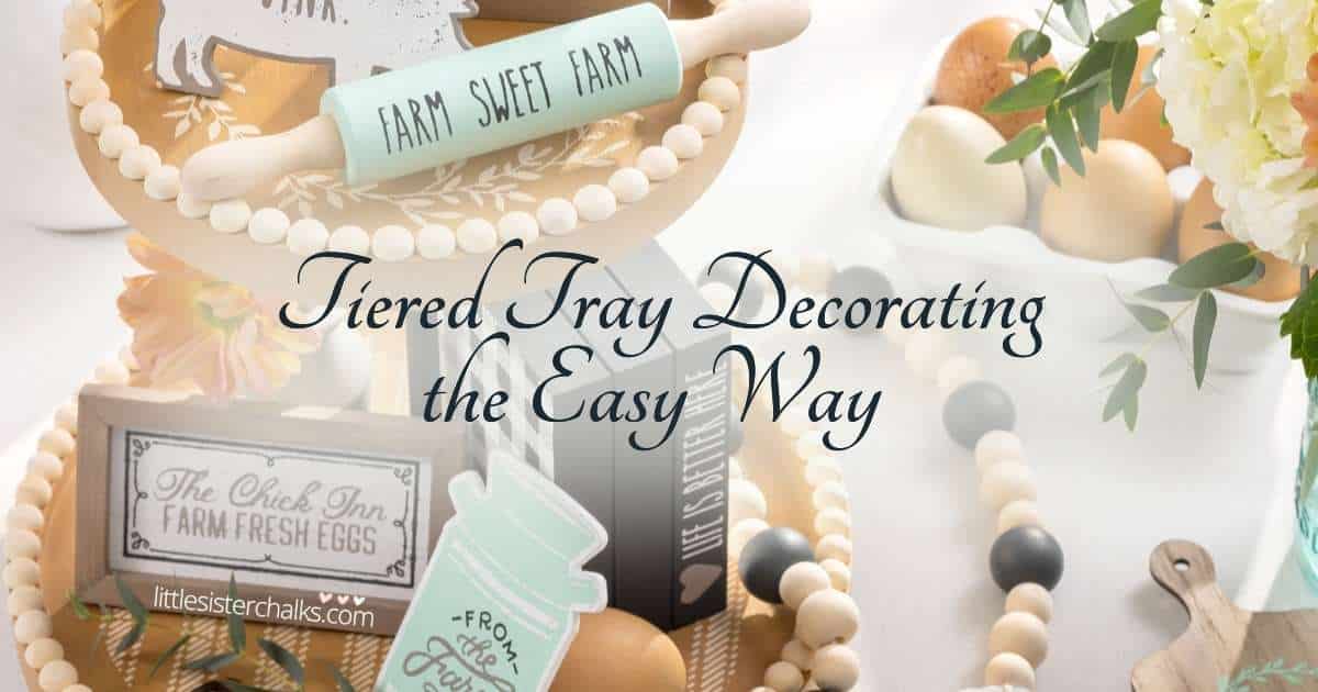 Close-up picture of a farmhouse tiered tray with a mini rolling pin, mini book stack, milk can cutout, mini box frame with a filled egg carton to the right and farmhouse beads with the words Tiered Tray Decorating the Easy Way