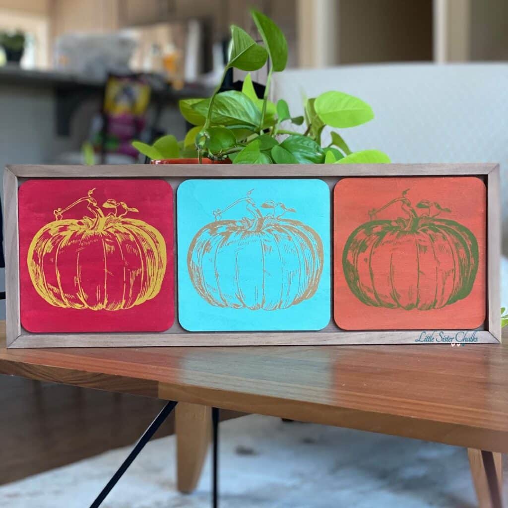 A box frame sitting on a coffee table with a pothos plant behind it with 3 pumpkins chalked in different colors, from left to right: red background/yellow pumpkin, blue background/gold pumpkin, and orange background/green pumpkin.