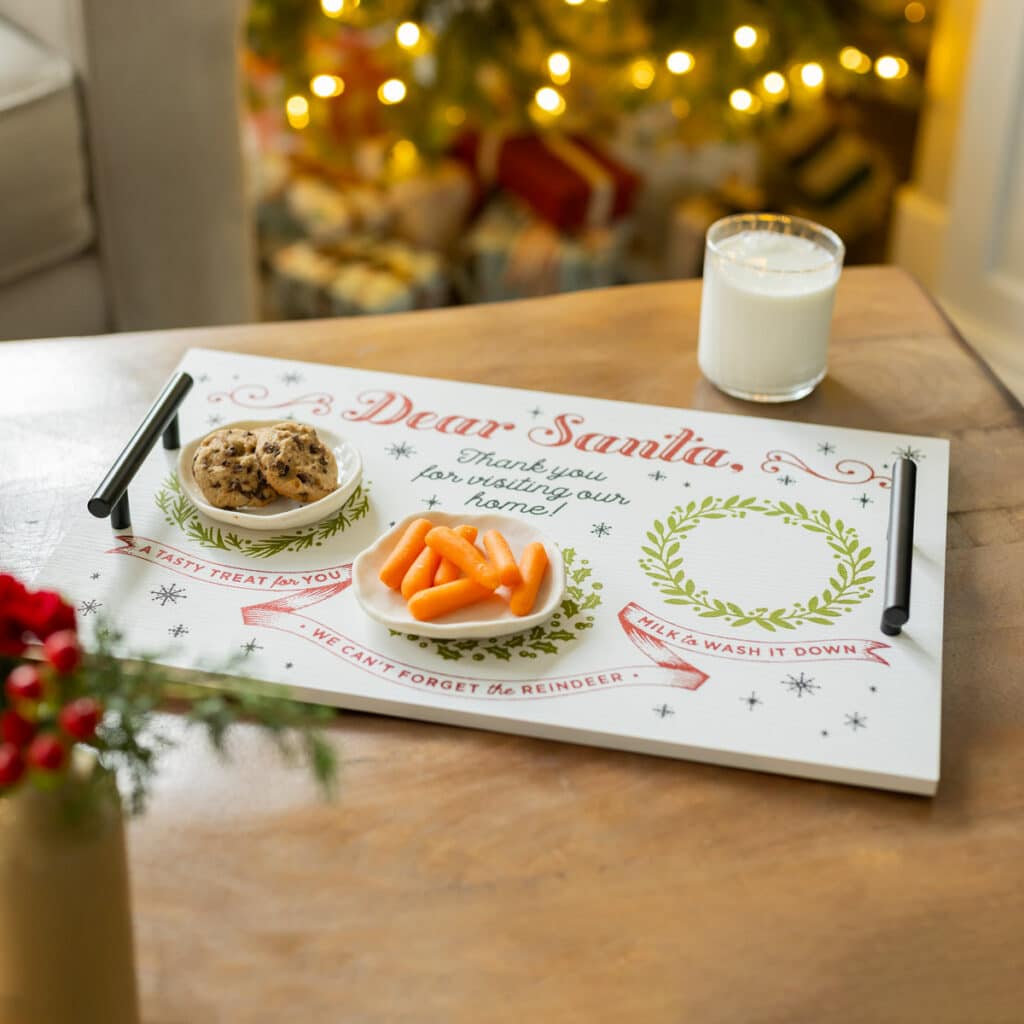 The finished Santa Tray ChalkMade Kit project on a coffee table flanked by a glass of milk with the words "Dear Santa, thank you for visiting our home! A tasty treat for you. We can't forget thee reindeer. Mile to wash it down." with two small plates on the tray holding chocolate chip cookies and carrots for the blog post "Chalk your own Santa Tray the Easy Way".