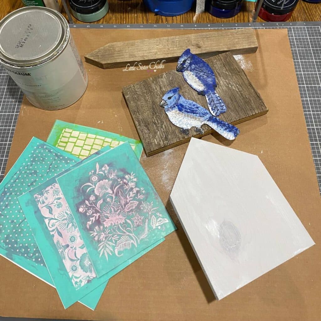A table top with a white house cut from wood, stencils, a piece or scrap wood with two blue birds painted blue and a can of paint for the blog post "DIY Scrap Wood Projects".