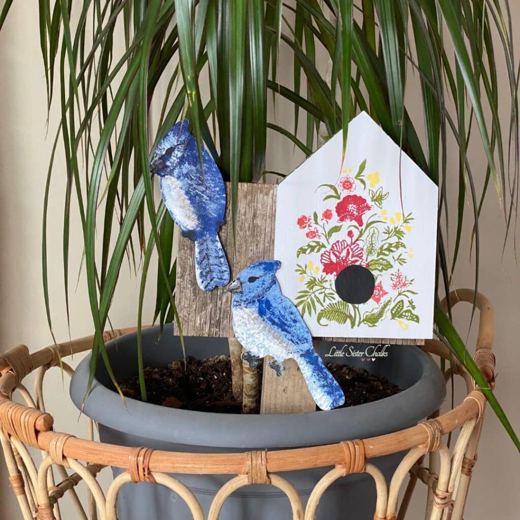A potted plan with a garden stake make of scrap wood, a white bird house chalked with red and yellow flowers, and 2 painted blue birds for the blog post "DIY Scrap Wood Projects".