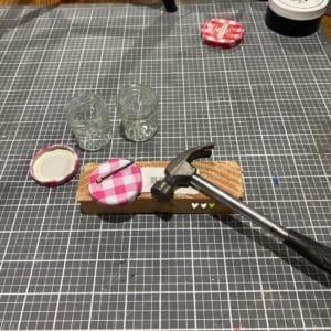 Two empty mini jam jars, two pink and white lids, a nail, a hammer and a piece of wood on a craft table for the blog post "Mini Jam Jar Craft Ideas".