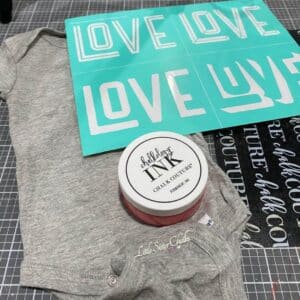 A photo for supplies to decorate a grey onesie with Chalk Couture including a jar of Chalkology Ink, an ink mat, and the Chalk Couture Retro Love stencil for the blog post "Use Chalk Couture Ink to Wear Your Art".