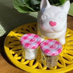 A white ceramic plant container shaped like a dog with a pink heart nose and a set of mini salt and pepper shakers made from mini jam jars with pink and white lids sitting on a yellow sunflower trivet for the blog post "Mini Jam Jar Craft Ideas".