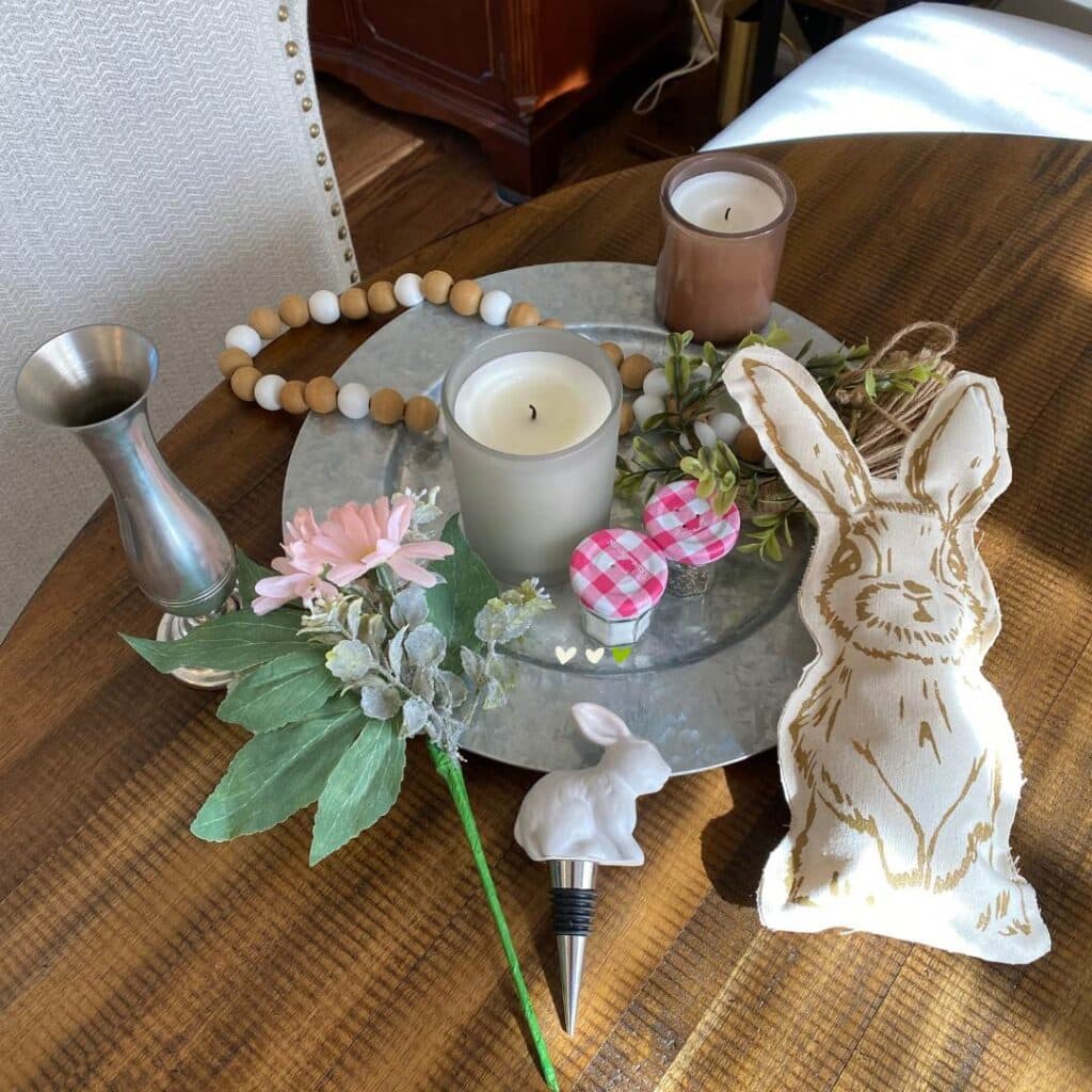Supplies to make a table center piece including a silver charger plate, a stuffed bunny, fake pink flowers and greenery, a white bunny wine stopper, a silver vase, a bead garland, two candles and a 2 mini jam jars with pink and white plaid lids for the blog post "Nine Ways to Use a DIY Stuffed Bunny in Your Decor this Spring".
