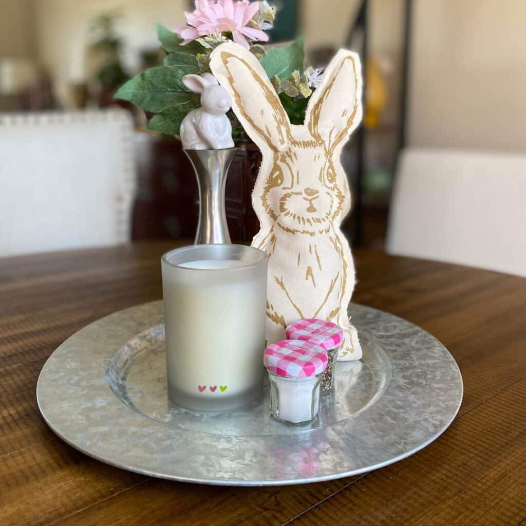 A table centerpiece including a stuffed bunny, 2 mini jam jars with pink and white plaid lids, a white candle and a white bunny wine stopper with some pink flowers and greenery in a silver vase all on a silver charger plate for the blog post "Nine Ways to Use a DIY Stuffed Bunny in Your Decor this Spring".