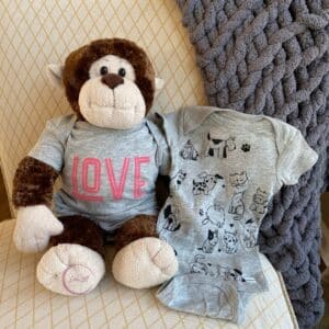 A photo of a stuffed monkey sitting on a cream chair with a gold diamond pattern wearing a grey onesie with the word Love inked on it with Chalk Couture Chalkology Ink in Couture Coral holding another grey onesie with the animals from the Chalk Couture Fur Baby stencil inked in Black Velvet Chalkology Ink for the blog post "Use Chalk Couture Ink to Wear Your Art".