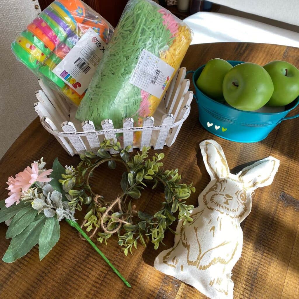 Supplies to make a basket including a stuffed bunny, fake pink flowers and greenery, a white picket fence basket, a teal blue metal tub, fake grass in pink, yellow and green, plastic Easter eggs, and 3 fake green apples on a table for the blog post "Nine Ways to Use a DIY Stuffed Bunny in Your Decor this Spring".