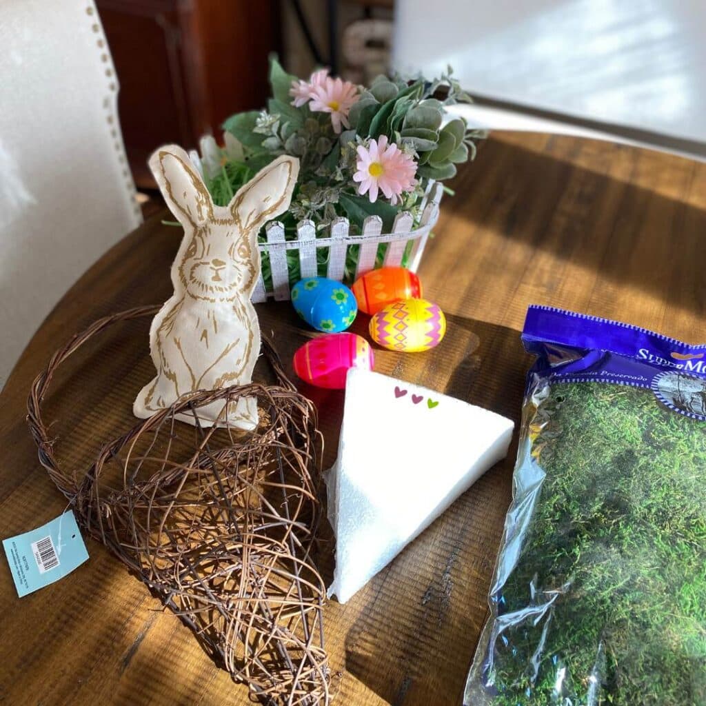 Supplies to make a door hanger including a stuffed bunny, a white styrofoam triangle, moss, plastic easter eggs, a white basket, fake pink flowers and greenery and a grapevine hanger for the blog post "Nine Ways to Use a DIY Stuffed Bunny in Your Decor this Spring".