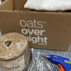 A cardboard box, an off-white tea towel, a roll of twine, an exacto knife, glue sticks and a glue gun on a craft mat for the blog post "Two Simple Ways to Reuse Cardboard Boxes Around Your Home".