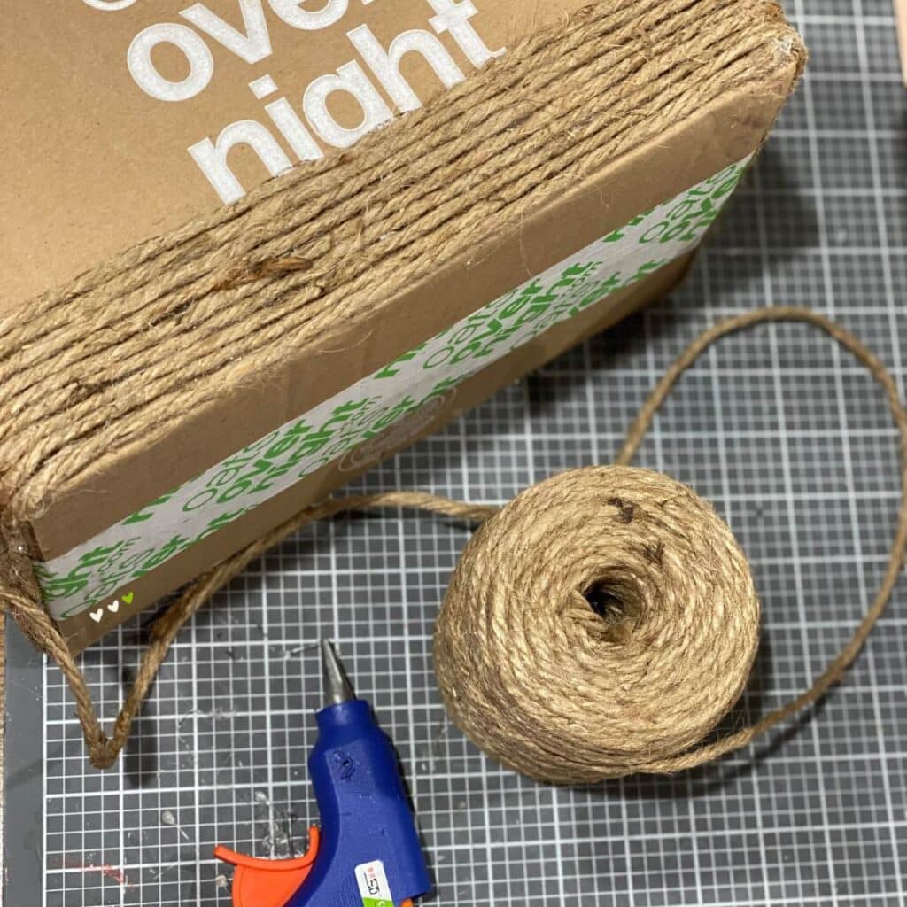 A photo of a cardboard box being wrapped in twine with a hot glue gun on a craft mat for the blog post "Two Simple Ways to Reuse Cardboard Boxes Around Your Home".