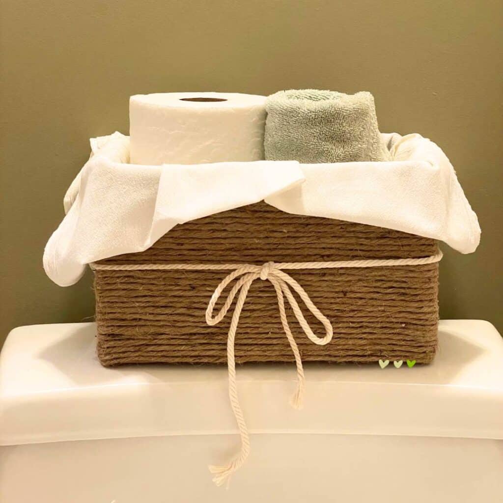 A basket made from a card board box and twine with an off-white tea towel liner and an off-white cord tied in a bow around the basket holding toilet paper and a blue rolled hand towel on top of a white commode for the blog post "Two Simple Ways to Reuse Cardboard Boxes Around Your Home".
