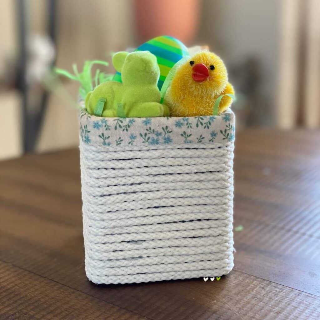 A small basket on a tabletop made from a box and off-white cord with a ribbon with little blue flower surrounding the rim holding green Easter grass, a blue and green plastic Easter egg, a small yellow chick and a small green bunny for the blog post "Two Simple Ways to Reuse Cardboard Boxes Around Your Home".