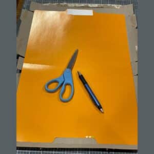 An orange file folder with a pair of scissors with blue handles, and a blue pen sitting on top over a flattened cereal box on a craft mat for the blog post "Two Simple Ways to Reuse Cardboard Boxes Around Your Home".