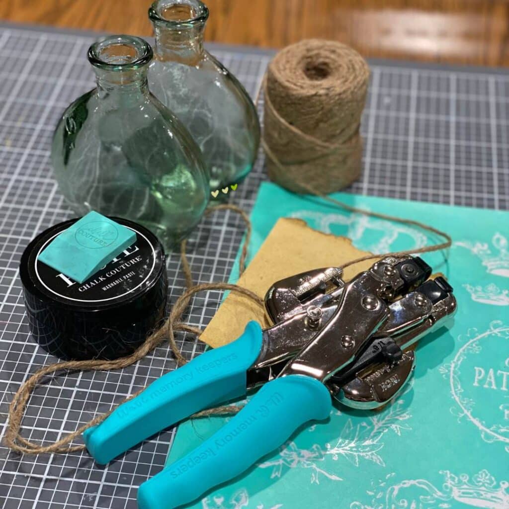 Two glass bottles, a jar of black chalk paste, a squeegee, some twine, a scrap piece of paper, a hole punch and a Chalk Couture stencil on a craft mat for the blog post "Thrift Flips: 5 Projects in 5 Days for an Earth Day Challenge".