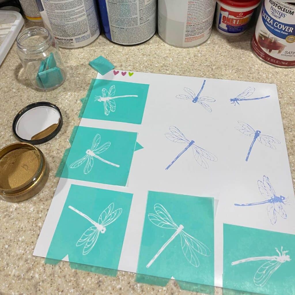 A Chalk Couture white magnetic sheet with dragonflies chalked in blue and 5 dragonfly stencils applied flanked by a jar of gold chalk paste on a counter for the blog post "How to Upcycle an Old Cookie Sheet with Chalk Couture".