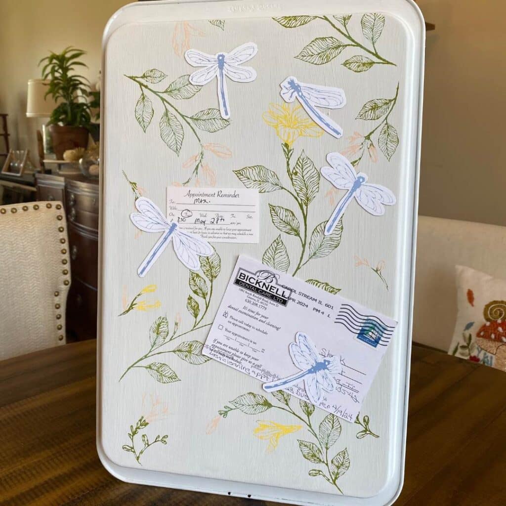 A memo board made from an old cookie sheet decorated with the Chalk Couture Dragonfly Vines stencil with two appointment cards held by blue dragonfly magnets for the blog post "How to Upcycle an Old Cookie Sheet with Chalk Couture".