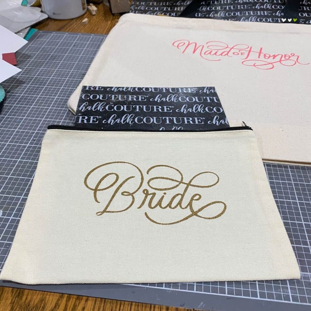 A canvas tote with "Maid of Honor" stenciled in coral ink and a canvas zippered bag with "Bride" stenciled in gold ink on a craft mat for the blog post "Customize Your Hats & Totes with Chalk Couture Ink".