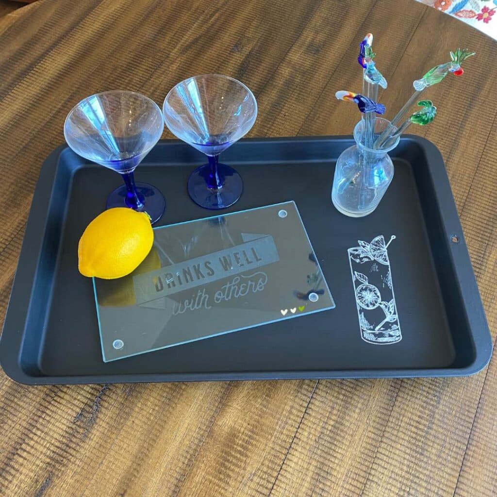 A cookie sheet upcycled into a serving tray styled with an etched cutting board that says "Drinks Well with Others", a lemon, two glasses, glass stir stick with birds and a palm tree for the blog post "How to Upcycle an Old Cookie Sheet with Chalk Couture".