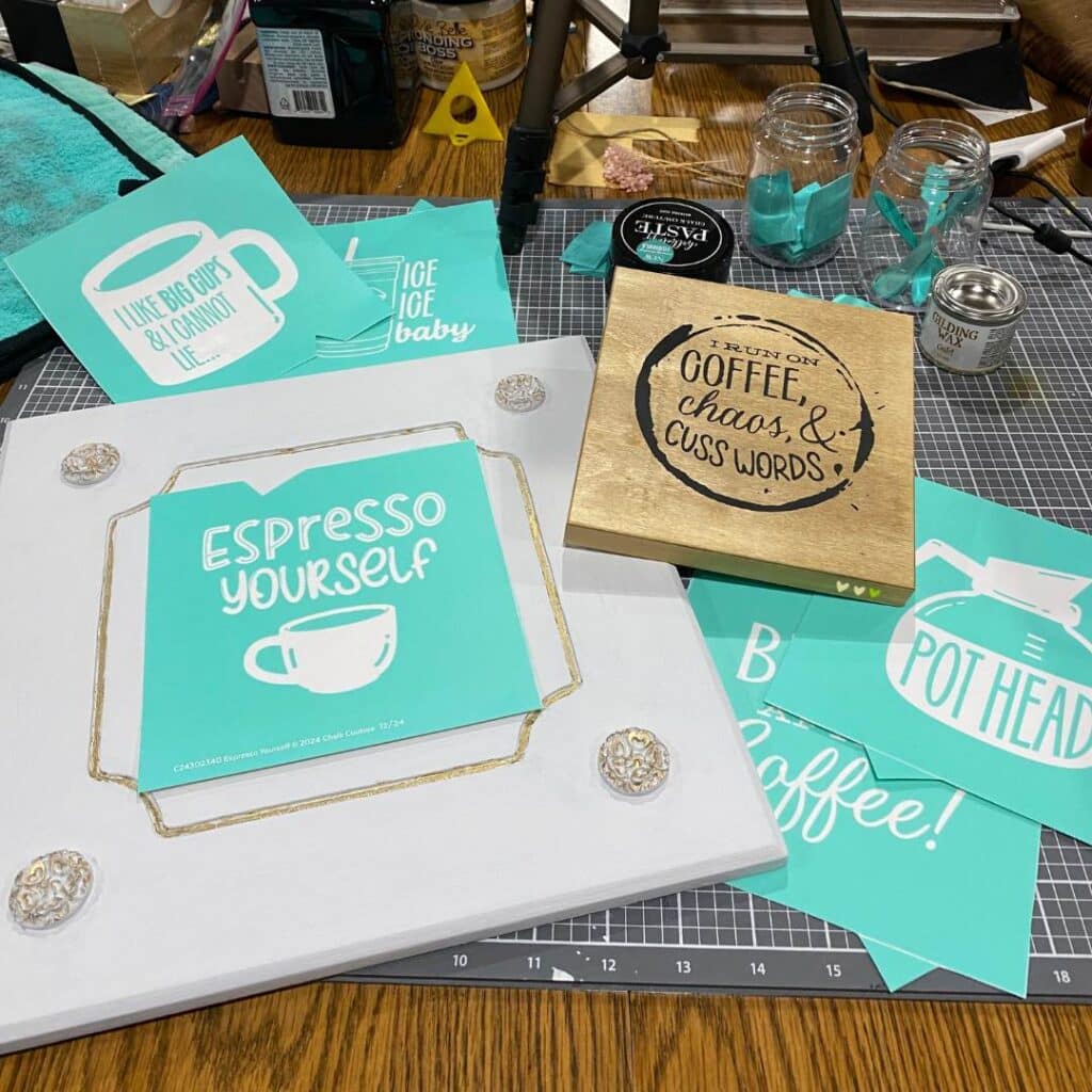 A white wood surface with a stencil that says "Espresso Yourself" laying on top of it flanked by a small wood sign with the words "Coffee, Chasos & Cuss Words", other coffee related stencils, black chalk paste, a jar of squeegess and a jar of stir sticks on a craft mat for the blog post "Thrift Flips: 5 Projects in 5 Days for an Earth Day Challenge".