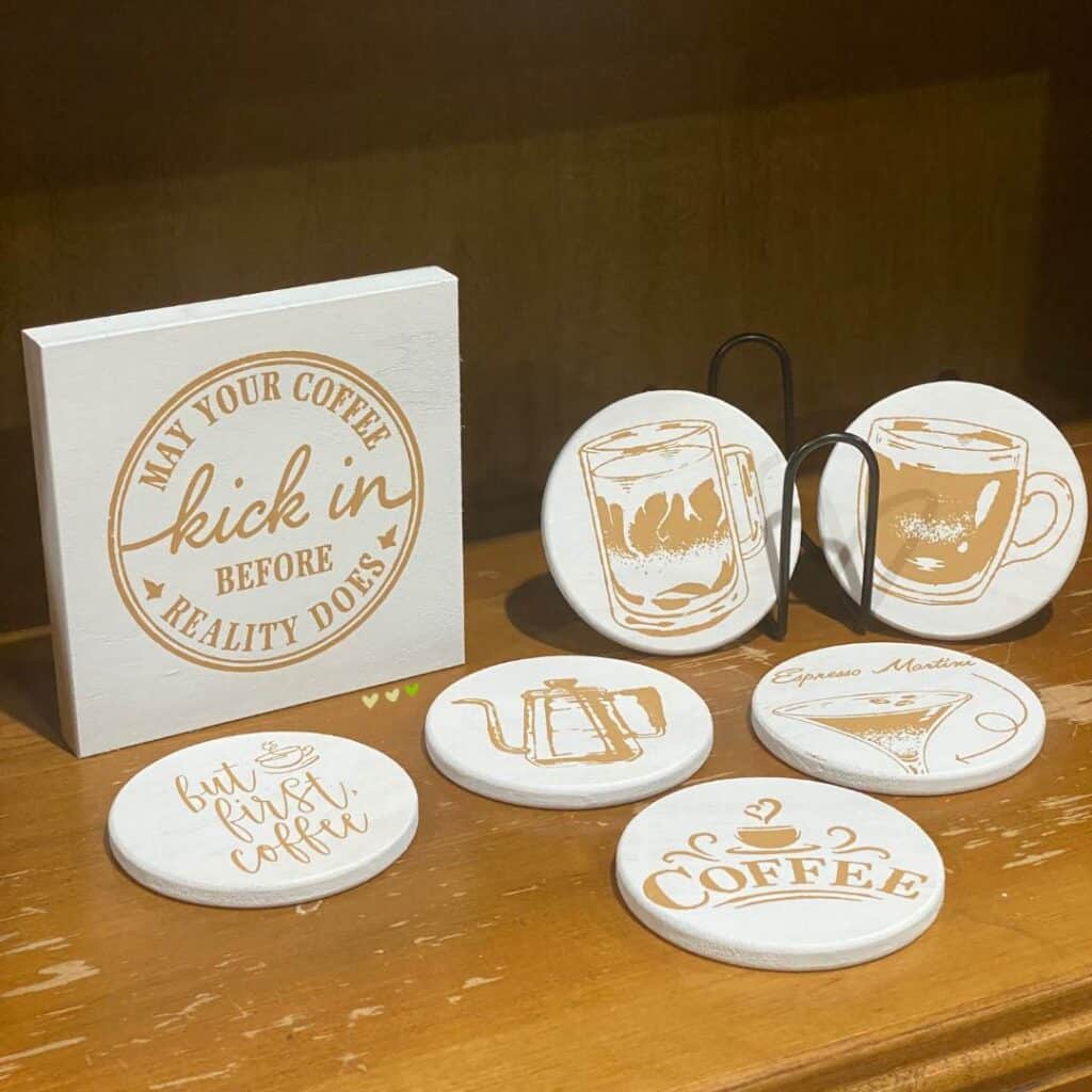 A set of 6 white wood coasters and a white wood box frame in coffee designs done in camel chalk paste. Three of the coasters have coffee images, one has a coffee pot, one has the word "coffee" with a cup above it and the last one has the words "but first, coffee" with a cup. The box frame has a circle with the words "May Your Coffee Kick In Before Reality Does" within it for the blog post "Bar Cart Decor Ideas that will have You Raising Your Glass".