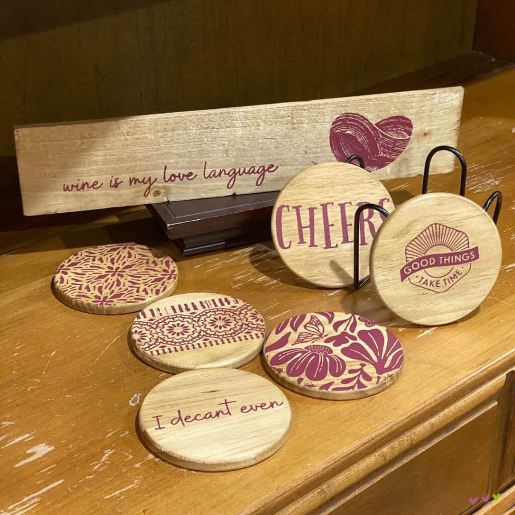A set of six wooden coaters and a small sign with wine designs done in red wine colored chalk paste. Three coasters have patterns, one coaster has the word "Cheers", one coaster has the words "I decant even", and the last coaster has the words "Good Things Take Time"; the sign says "wine is my love language" flanked by a heart for the blog post "Bar Cart Decor Ideas that will have You Raising Your Glass".