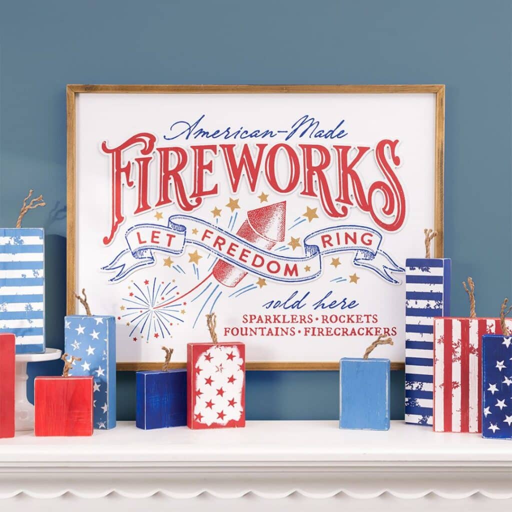 A white wood sign using the Chalk Couture "Fireworks Sold Here" stencil done in red, blue and gold hanging above a mantel with a variety of wood block firecrackers in blue and red decorated in starats and stripes for the blog post "Fly the Red, White & Blue with the 2024 Patriotic Collection from Chalk Couture".