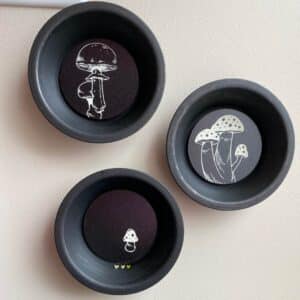 Three mini tart pans repurposed into wall art by painting them with black craft paint and inserting Chalk Couture Chalkable Chips with white stenciled mushrooms for the blog post "Simple Baking Pan Crafts You Can Use to Repurpose Your Old Bakeware".