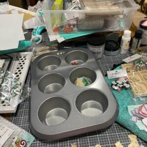 A muffin tin on a craft table surrounded by small Christmas embellishments like bottle brush trees, stickers, labels and small wooden snowflakes for the blog post "Simple Baking Pan Crafts You Can Use to Repurpose Your Old Bakeware".