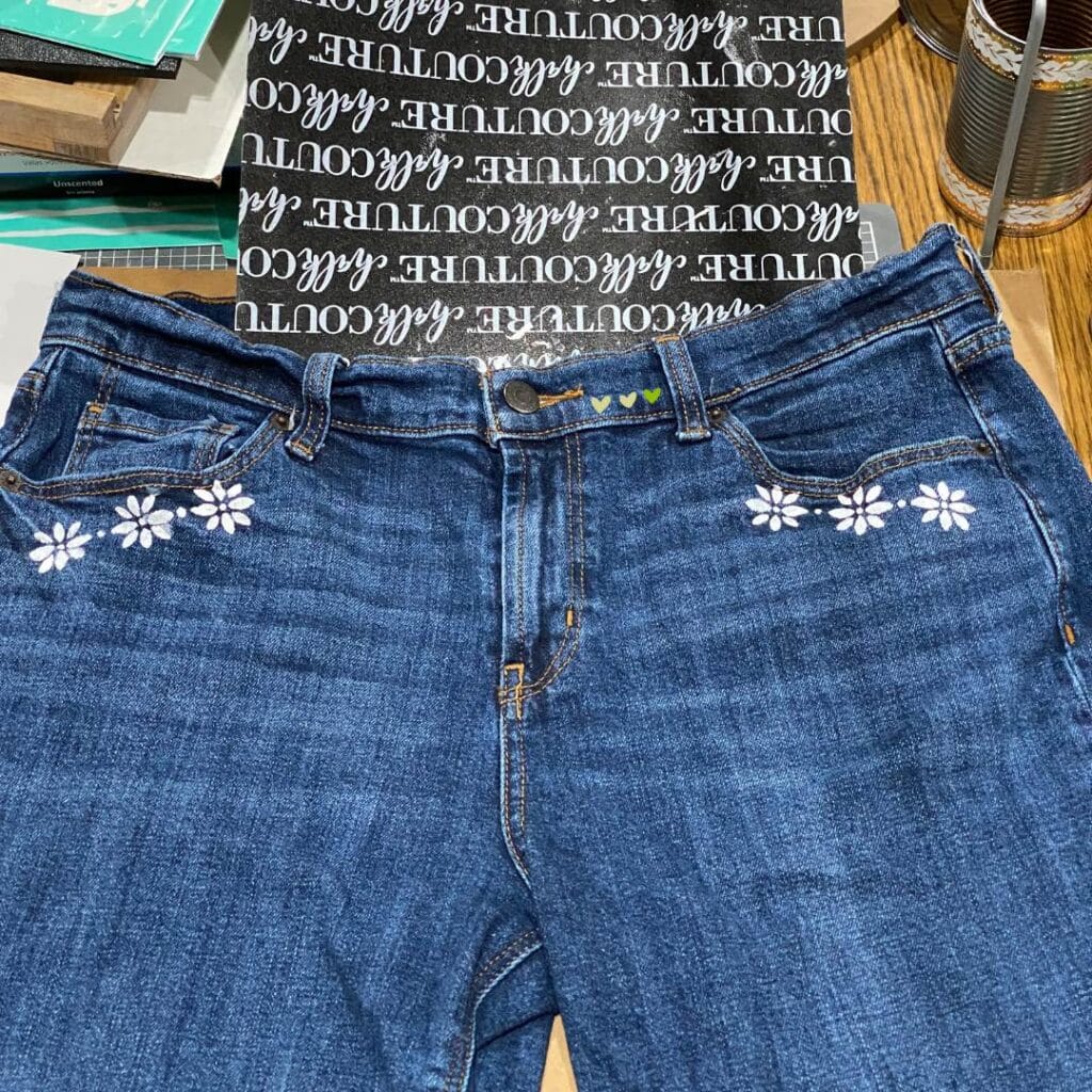A photo of a pair of jeans on a craft table with small flowers inked in white under the front pockets for the blog post "Stencil & Stitch Your Style with Chalk Couture: Upcycled Bottoms Edition".