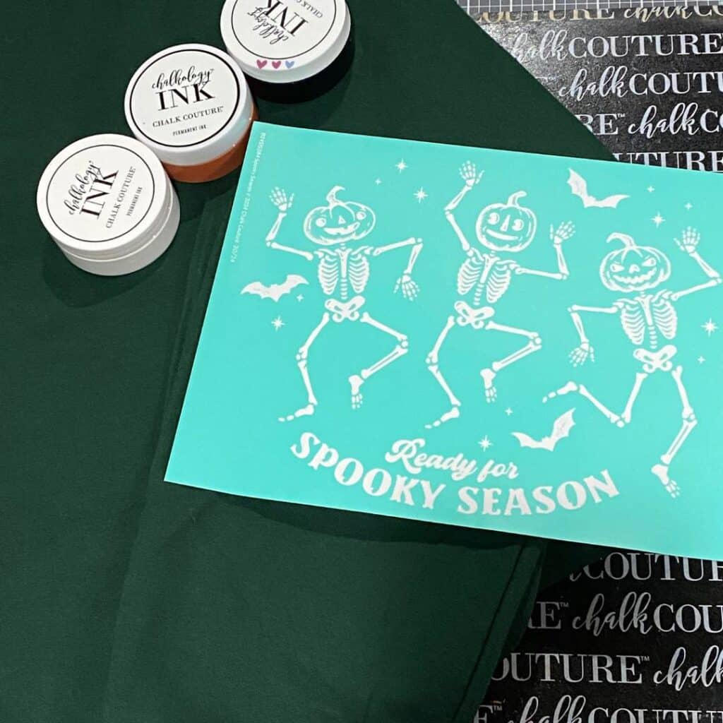 A forest green sweatshirt, an ink mat, a Chalk Couture stencil with 3 skeletons with pumpkin heads, and 3 jars of Chalkology ink in orange, white and black on a craft table for the blog post "Stencil & Stitch Your Style: Upcycled Outerwear Edition".