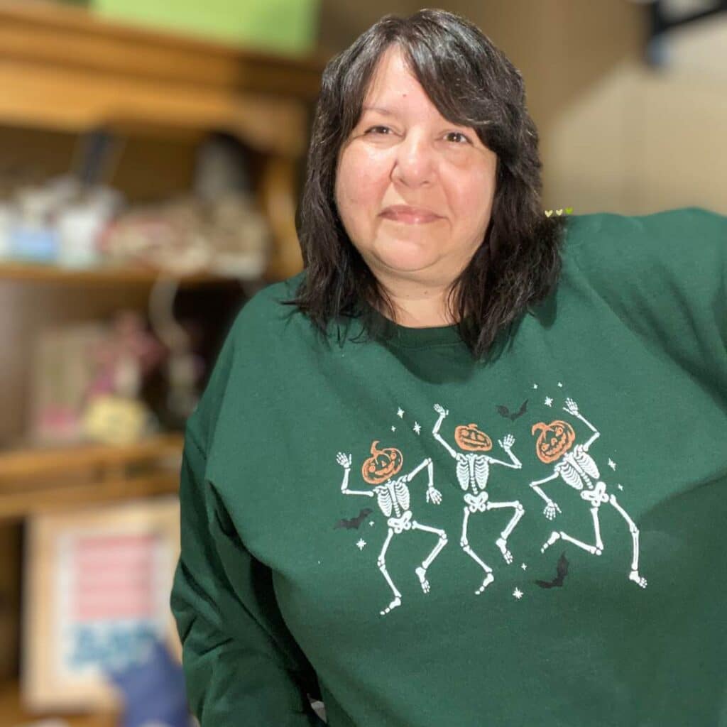 A photo of Luana Slenk, Chalk Couture Independent Designer, wearing a forest green sweatshirt with a stenciled design including 3 skeletons with pumpkin heads, bats, and stars for the blog post "Stencil & Stitch Your Style: Upcycled Outerwear Edition".