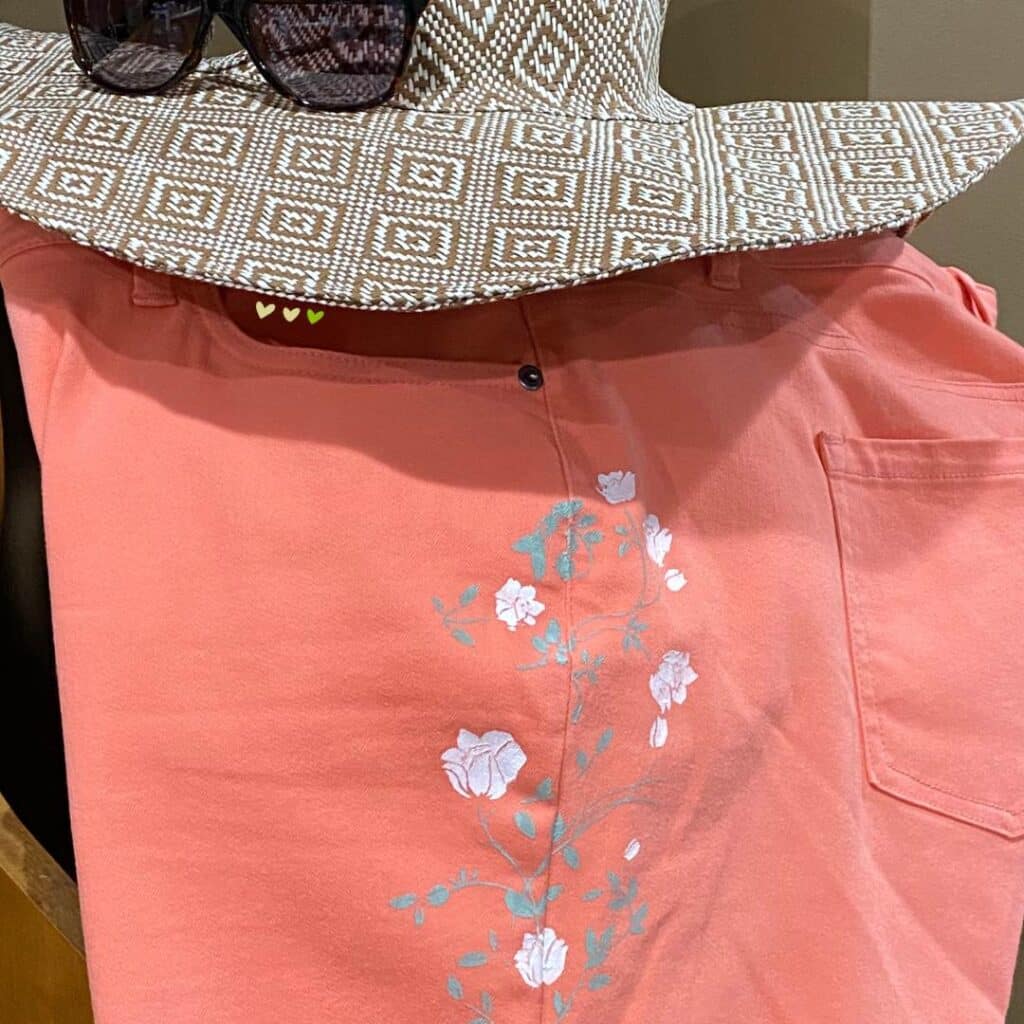 A photo of a the leg of a pair of peach capri pants with a stencil design of white flowers and green stems paired with a sun hat and sunglasses for the blog post "Stencil & Stitch Your Style with Chalk Couture: Upcycled Bottoms Edition".