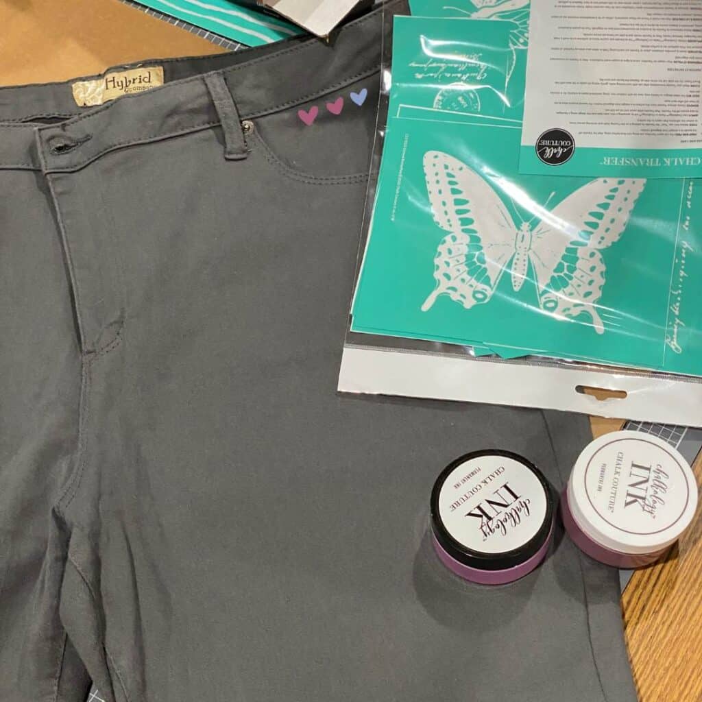 A pair of grey Bermuda shorts on a craft table with a butterfly stencil and two jars of Chalk Couture Chalkology Ink in Lilac and Fig for the blog post "Stencil & Stitch Your Style with Chalk Couture: Upcycled Bottoms Edition".