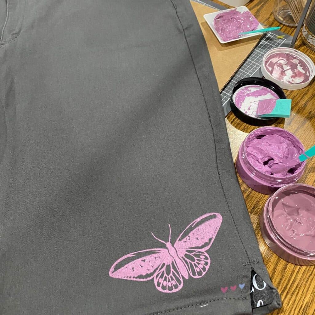 A pair of grey Bermuda shorts with a butterfly etched near the hem of one leg with a jar of Lilac and a jar of Fig Chalk Couture Chalkolgoy Ink for the blog post "Stencil & Stitch Your Style with Chalk Couture: Upcycled Bottoms Edition".