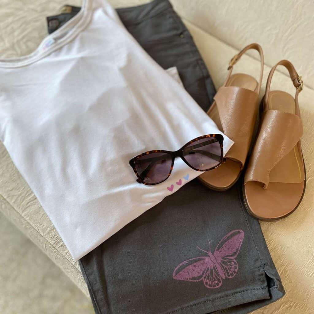 A pair of grey Bermuda shorts with a butterfly stenciled in purple paired with a white shirt, sunglasses and a pair of tan sandals for the blog post "Stencil & Stitch Your Style with Chalk Couture: Upcycled Bottoms Edition".