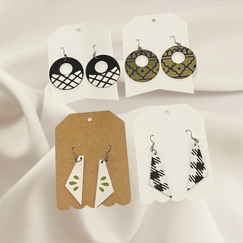 A photo of 4 pairs of wooden earrings laying on a white fabric background decorated with Chalk Couture products - 2 round pairs of earrings: one done in black and white graphic design and one with a green background and shimmer blue boho design, 2 pairs of tapered pendants one with a black and white graphic design and one with a white background and a small green design for the blog post "Elevate Your Style with Wearable Art Earrings featuring Chalk Couture".
