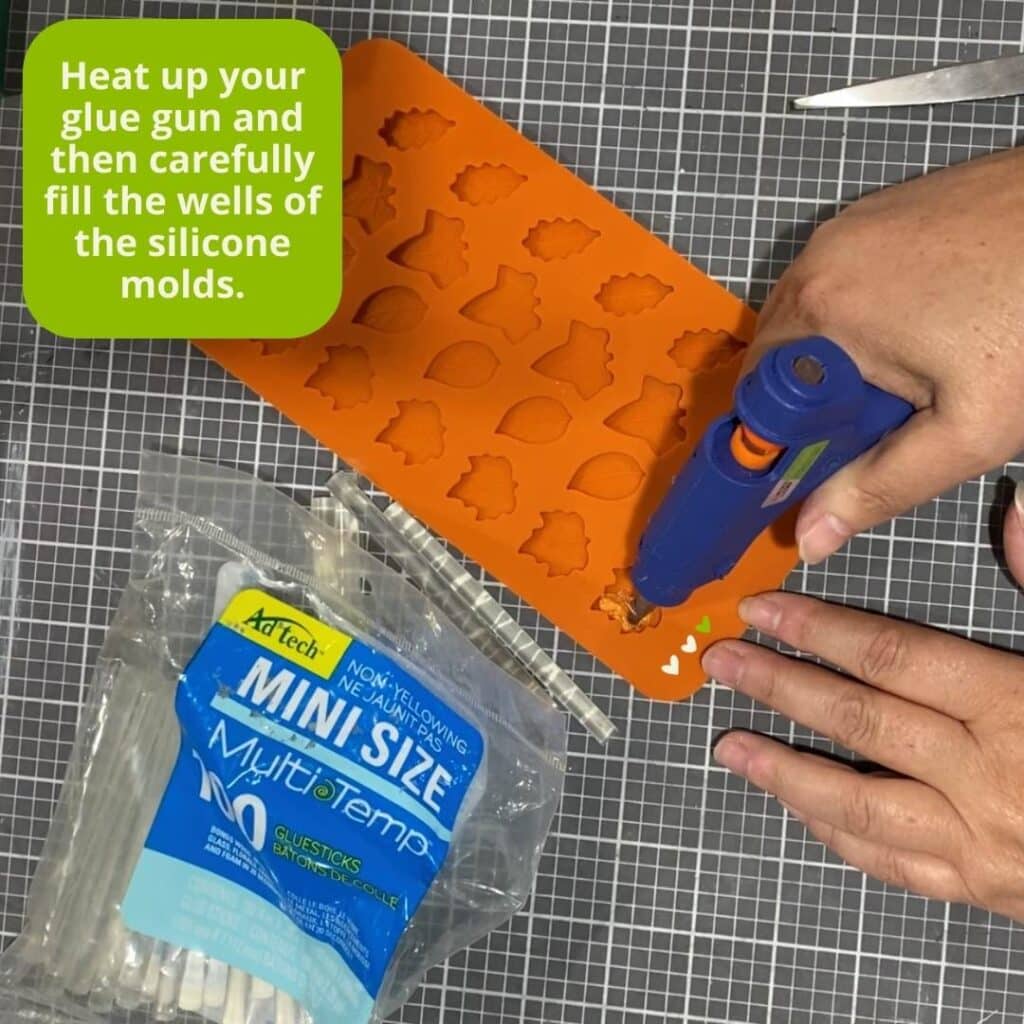 A photo of a hand holding a glue gun on a craft mat, an orange silicone leaf mold and mini glue sticks with instructions to "heat up your glue gun and then carefully fill the wells of the silicone molds" for the blog "Transform Your Crafts with the Perfect Finish: DIY Molded Hot Glue Embellishments".