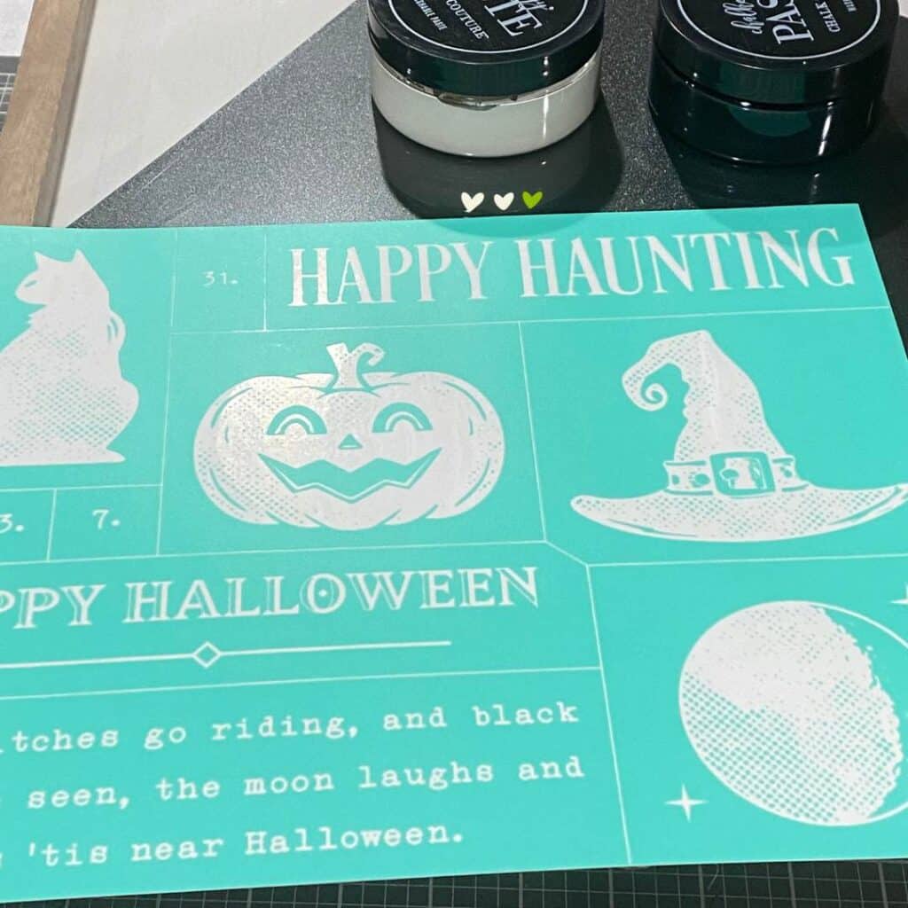 Chalk Couture supplies on a craft table including the Happy Haunting stencil, an Outside the Box Frame and a black insert, and one jar of black paste and one jar of white paste for the blog post "Theme It Up for Halloween".