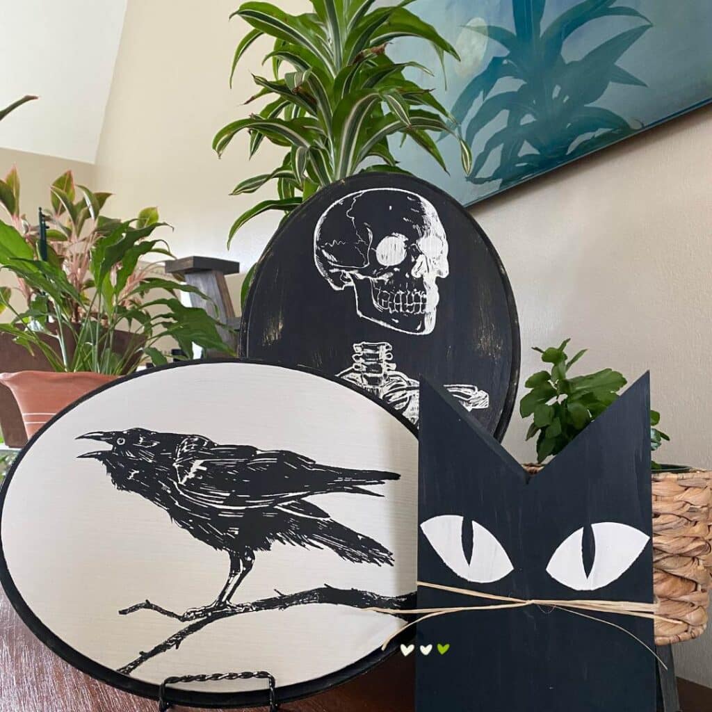 Three Halloween decor items including (from left to right) a black crow on an oval painted white with a black border, a white skeleton on a black oval, and a wooden black cat with white eyes and raffia for whiskers for the blog post "Theme It Up for Halloween".