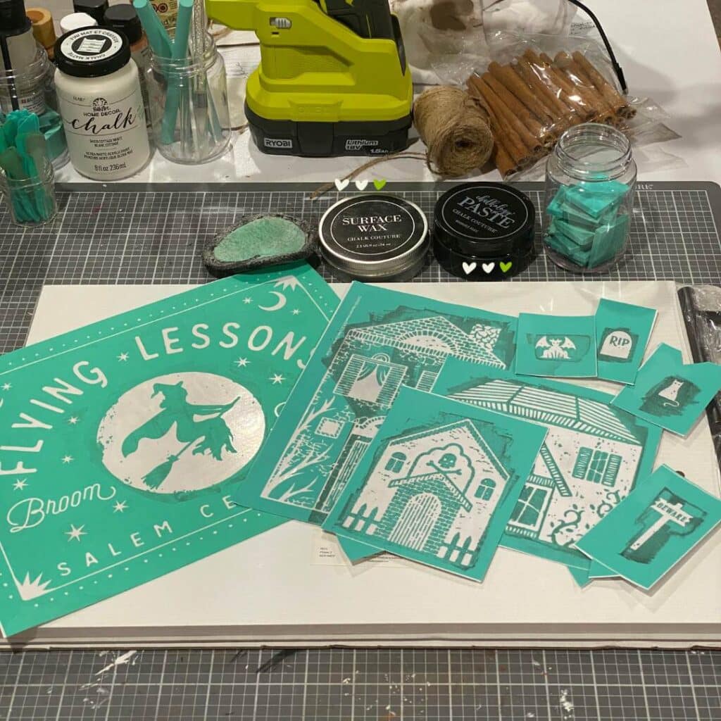A variety of craft supplies laying a craft table including stencils with a witch and spooky houses, a packaged white tray, Chalk Couture Black Velvet Chalkology Paste, squeegees in a jar, and surface wax and an applicator for the blog post "Theme It Up for Halloween".