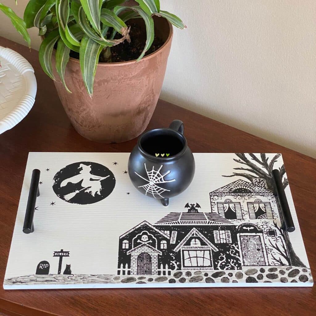 A white tray with a Halloween scene chalked in black chalk paste on a sideboard holding a cup that is shaped like a cauldron. They tray has a witch flying over spooky houses, a tree, a headstone, a black cat, a beware sign and cobblestone sidewalks for the blog post "Theme It Up for Halloween".