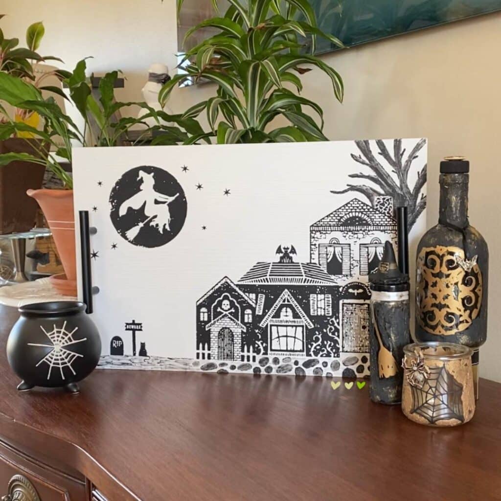 A white tray with a witch flying over spooky. houses chalked in black chalk paste flanked on the left by a black cup shaped like a cauldron with spider webs on it and on the right by 3 apothecary jars in black and gold for the blog post "Theme It Up for Halloween".