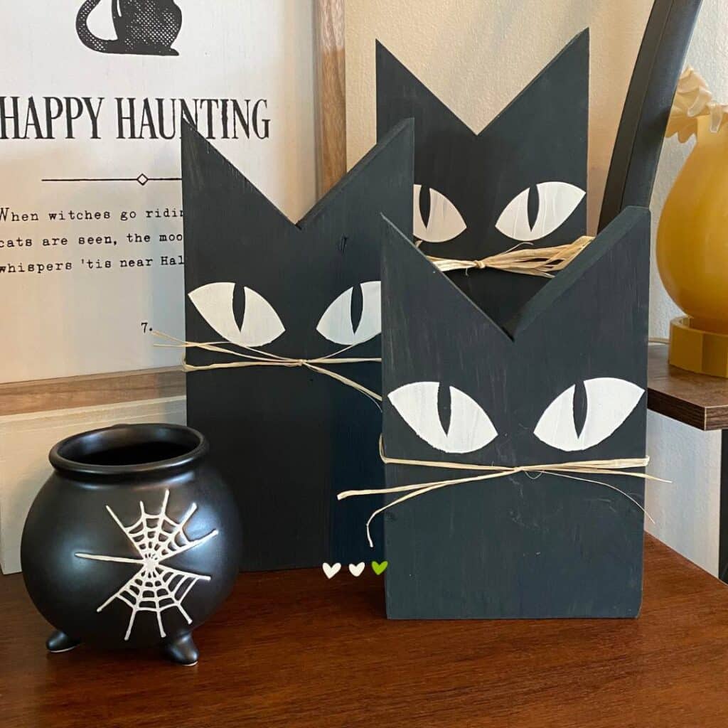 Three wooden black cats with white eyes and raffia for whiskers in sizes from 12" to 8" on a sideboard flanked by a black cauldron cup with white spider webs for the blog post "Theme It Up for Halloween".
