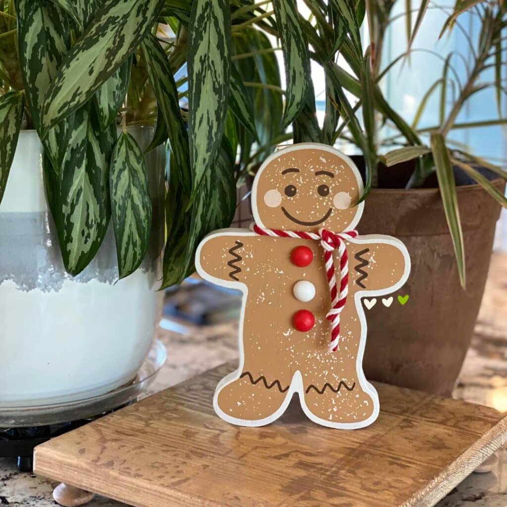 A gingerbread man shelf sitter sitting on a riser between two plants on a counter for the blog post "Christmas Shelf Decor".
